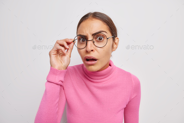 Photo of embarrassed brunette woman feels shocked stands speechless keeps hand on rim of spectacles