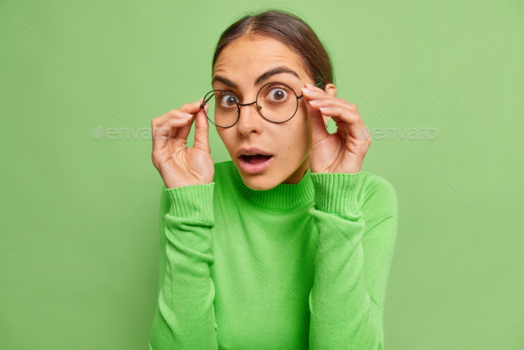 Photo of lovely impressed young woman keeps hands on rim of spectacles looks with great wonder keeps