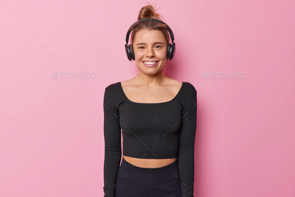 Waist up shot of active happy young sportswoman in black tracksuit listens favorite song via wireles