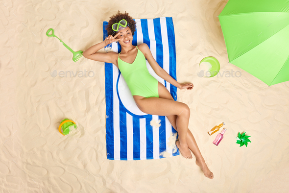 Overhead shot of cheerful curly haired woman wears snorkeling mask and green swimsuit makes peace ge