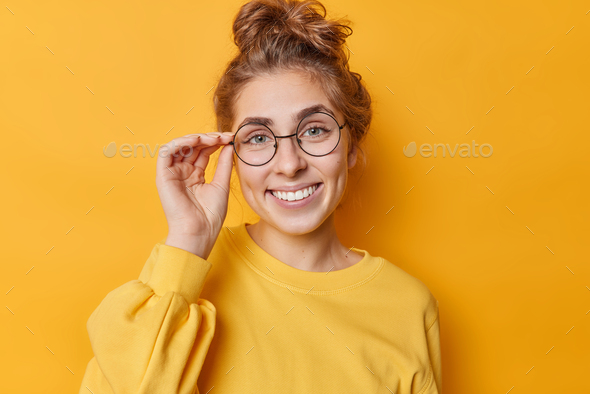Portrait of lovely European woman keeps hand on rim of spectacles smiles gladfully looks self confid