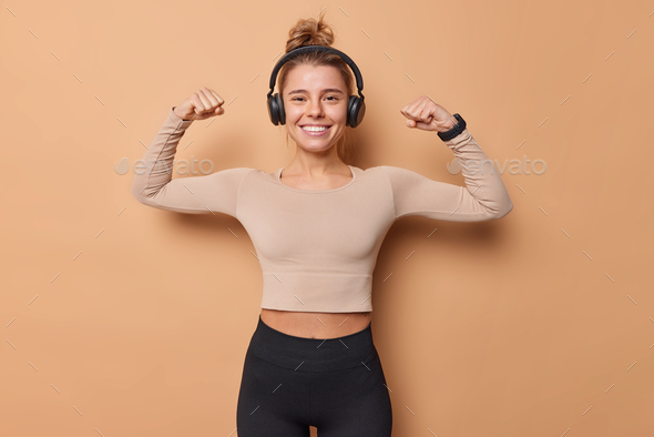 Horizontal shot of active slim young woman raises arms shows biceps after workout dressed in sportsw