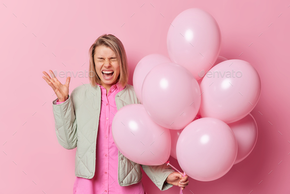 Emotional woman screams outraged shouts at someone holds bunch of inflated balloons keeps hand raise