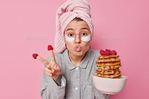 Funny young woman makes peace gesture keeps lips folded poses with delicious homemade pancakes appli
