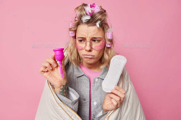 Upset young woman chooses between menstruation cup and sanitary napkin has bad mood during menstruat - Stock Photo - Images