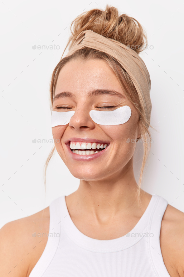 Vertical shot of cheerful woman smiles broadly applies beauty patches under eyes ro remove wrinkles
