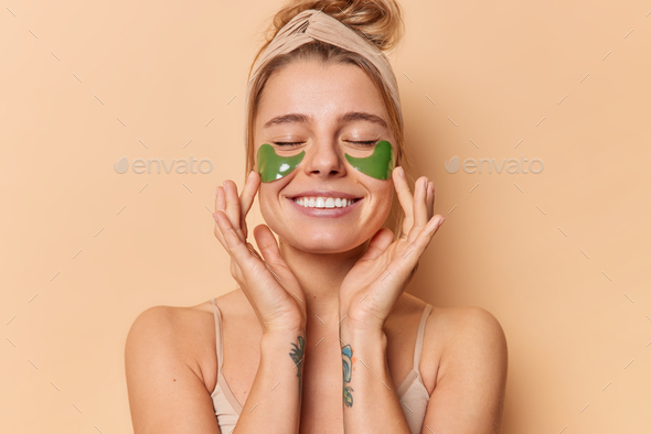 Pleased tender young woman applies green hydrogel patches under eyes to remove wrinkles wears headba
