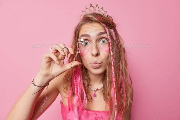 Surprised European woman prepares for party uses eyelashes curler applies makeup beauty hydrogel pat