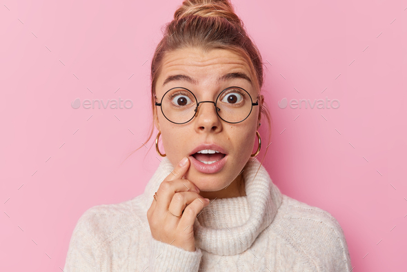 Scared woman stares shocked holds breath has mouth opened from amazement wears spectacles and sweate