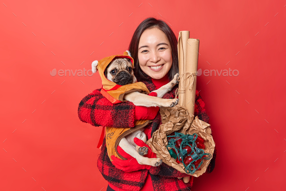 Horizontal shot of lovely cheerful dark haired Asian woman embraces pug dog dressed in winter clothe