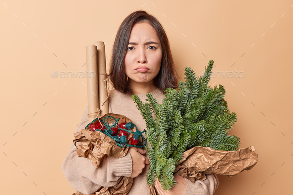 Upset disappointed Asian woman purses lips feels unhappy poses with spruce branches rolled paper and