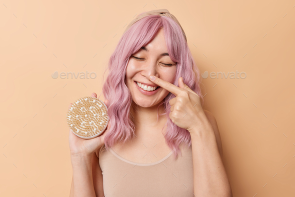 Horizontal shot of pink haired woman touches nose and smiles gently holds body brush has healthy ski