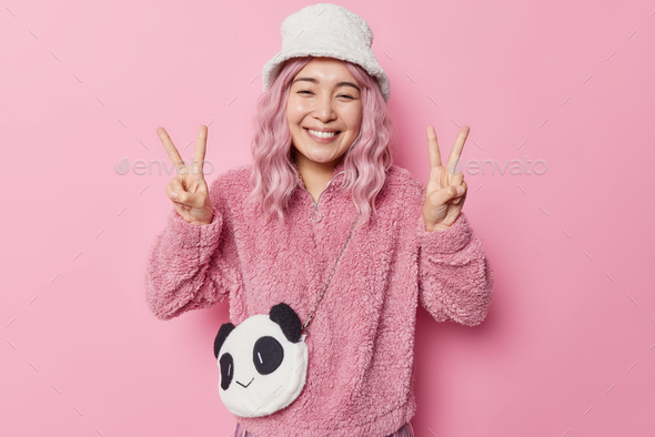 Smiling positive Asian woman makes peace gesture with both hands being in good mood enjoys life wear