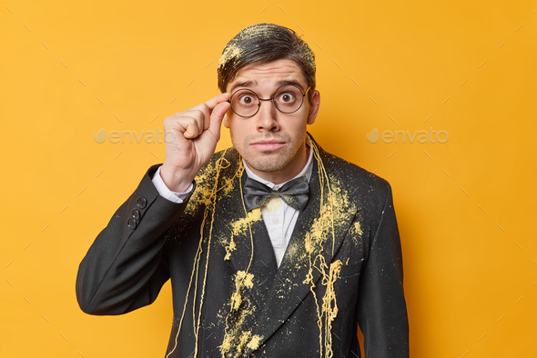 Impressed shocked man dressed in formal festive suit keeps hand on rim of spectacles smeared with se