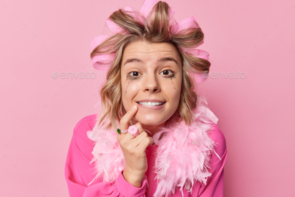 Headshot of cheerful woman with spoiled makeup applies hair curlers prepares for date wears festive