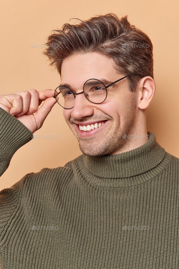 Curious positive adult European man keeps hand on rim of round spectacles smiles and concentrated aw