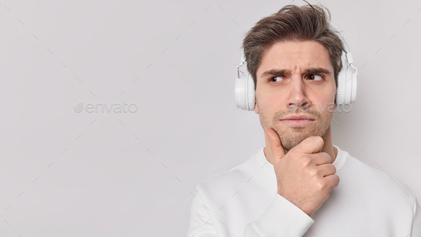 Serious male student listens educational content holds chin wears wireless headphones with good soun