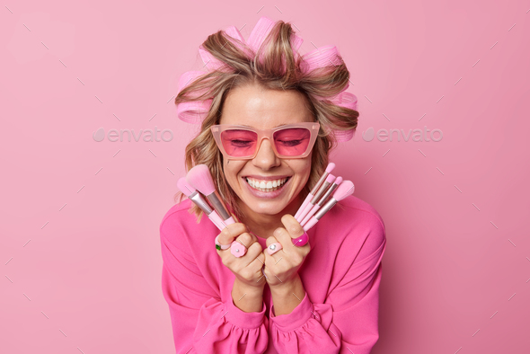 Happy lady applies hair rollers for making hairstyle wears trendy sunglasses sand blouse holds cosme