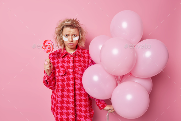 Frustrated upset woman has bad mood after party holds delcious round candy on stick and bunch of inf