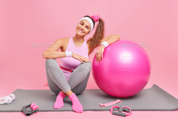 Horizontal shot of cheerful sportswoman leans on fitball does gymnastics at home dressed in sportswe