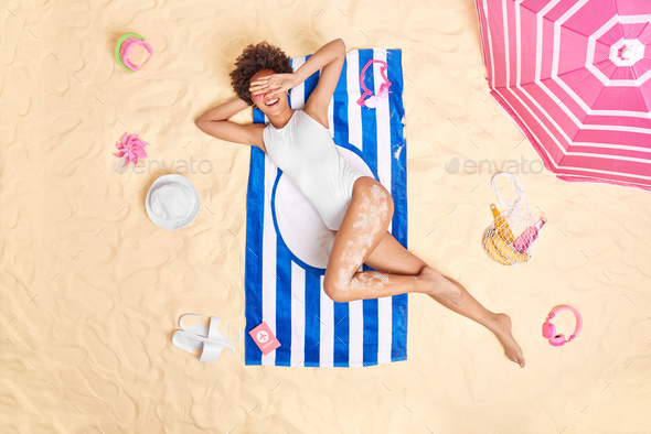 Cheerful young woman hides face with hand smiles gladfully wears white swimsuit poses on striped tow