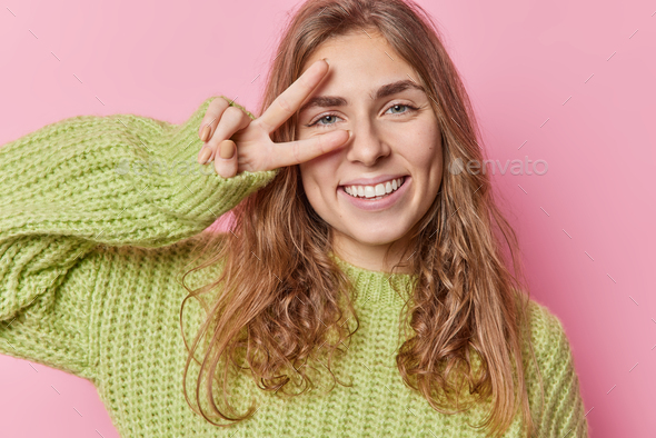 Positive peaceful blue eyed pretty woman with long wavy hair makes peace gesture over eye smiles gla