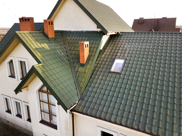 Green metal shingled house roof with attic plastic window and brick chimney.