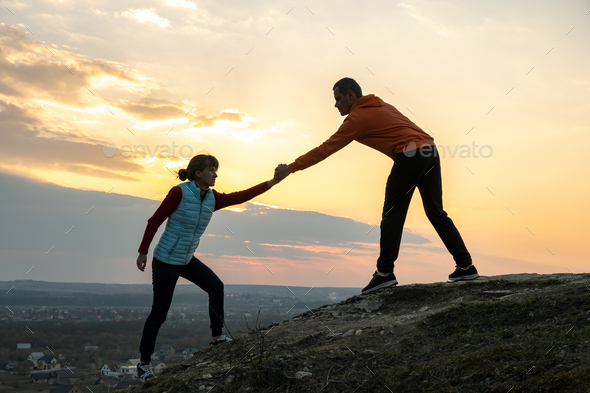 Man and woman hikers helping each other to climb stone at sunset in mountains.