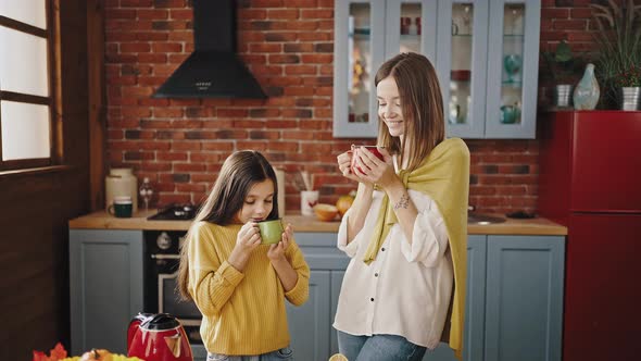 Young Mother and Her Little Daughter are Smiling Holding Colorful Cups with Tea and Inhaling It alt