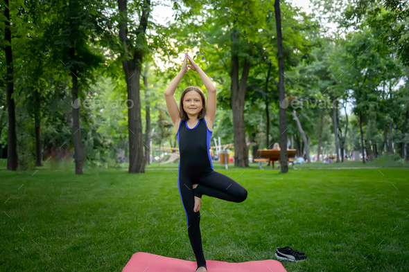 a little girl in the park is doing yoga, she stands on one leg Stock Photo  by romanchoknadii