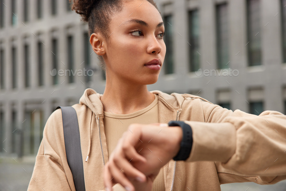 Horizontal shot of sporty woman dressed in brown hoodie wears wristwatch checks time looks seriously