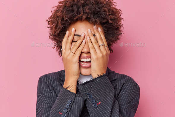 Happiness and emotions concept. Cheerful young curly woman hides face with hands giggles positively