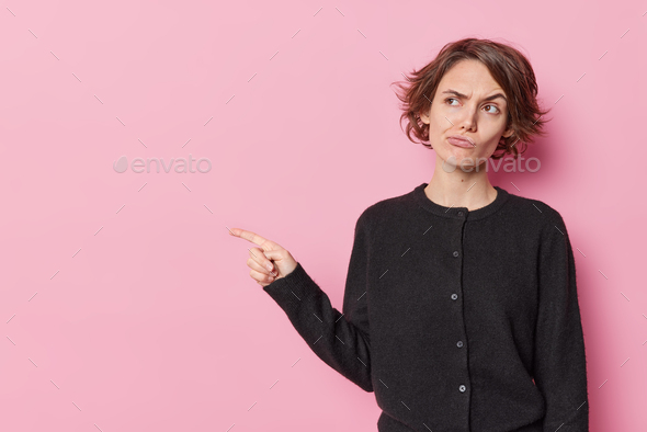 Horizontal shot of displeased woman with short hair raises eyebrows sulks face points index finger a