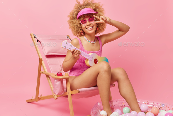 Positive carefree curly haired woman dressed in swimsuit makes peace gesture plays guitar being in g