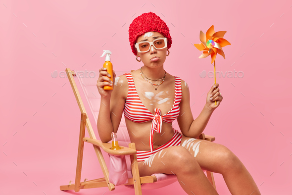 Sad woman with fit body has bad mood holds suncreen lotion and windmill  gets suntan at beach dressed Stock Photo by wayhomestudioo