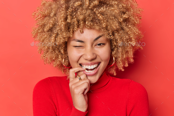 Positive curly haired young woman bites finger winks eye and smiles broadly shows white teeth being