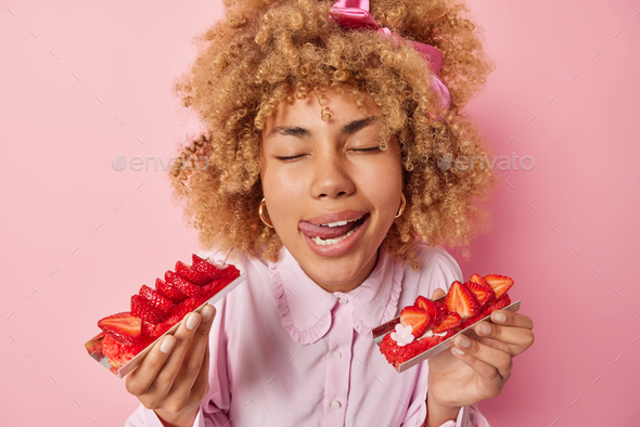 Photo of pleased curly haired woman licks lips with tongue gets pleasure from eating delicious straw