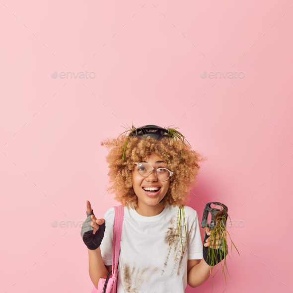 Positive active woman with curly hair wears protective helmet gloves spectacles dirty t shirt holds