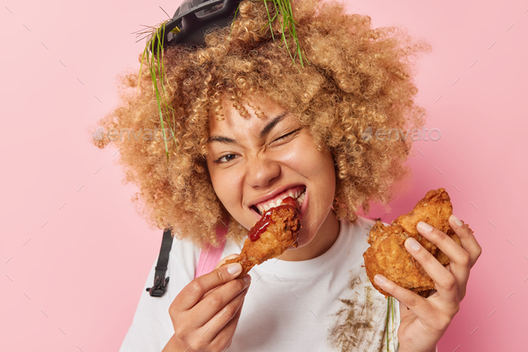 Headshot of curly haired woman eats appetizing fried nuggets with ketchup enjoys eating cheat meal w