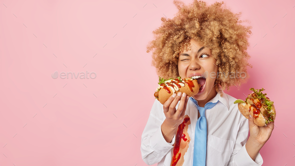 Unhealthy junk food and cheat meal concept. Curly haired woman eats delicious hot dog and hamburger