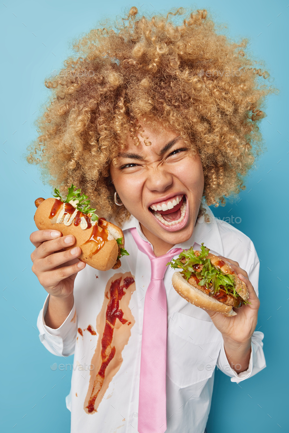 Curly haired woman enjoys eating cheat meal holds sandwich and hot dog enjoys favorite fast food exc