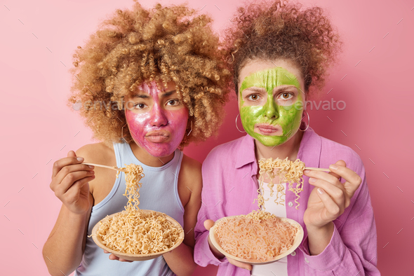 Serious women apply beauty mask on face eat delicious spaghetti frown face stand next to each other