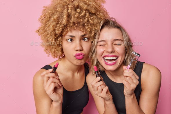 Two funny women foolish around apply lipstick use eyelashes curler put on makeup prepare for party w
