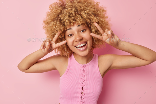 Positive curly haired woman makes peace gesture smiles happily at camera looks carefree enjoys life