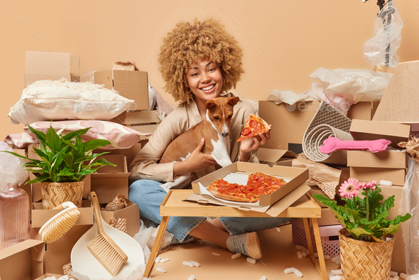 Good looking dark skinned African woman eats delicious pizza enjoys eating  fast food using mobile Stock Photo by wayhomestudioo