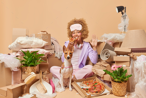 Indoor shot of happy woman eats pizza waves hello with palm sits crosed legs near dog surrounded by