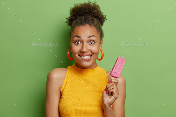 Horizontal shot of beautiful surprised woman bites lips holds appetizing ice cream with strawberry f