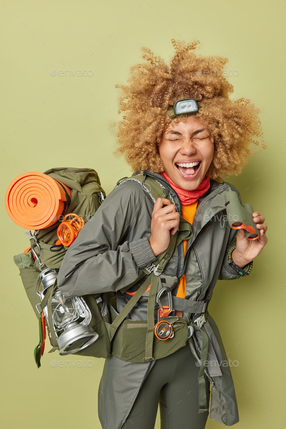 People travel adventure and recreation concept. Joyful curly haired woman holds binoculars laughs gl