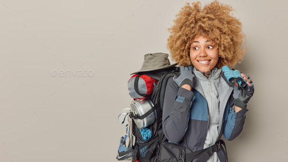 Positive female explorer uses binoculars to look into distance carries backpack has expedition dress