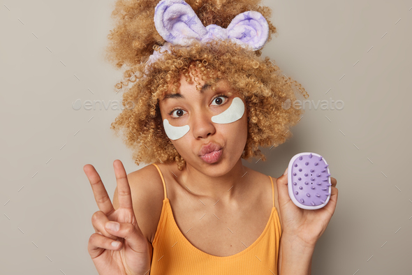 Indoor shot of curly haired young woman keeps lips folded makes peace gesture holds body brush wears